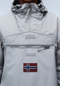 Lys grå, hættejakke med en lynlås-lomme, velcro-detaljer og et rektangulært mærke med det norske flag på forsiden.