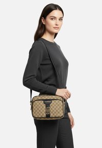 Borsa a tracolla color cuoio con motivo geometrico, dettagli neri, texture liscia e dettaglio con logo. Indossata da una modella con un outfit scuro.