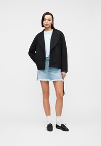 Jeune femme debout portant une veste noire, un pull bleu clair, une jupe en denim effilochée, des chaussettes blanches et des mocassins noirs sur un fond blanc.