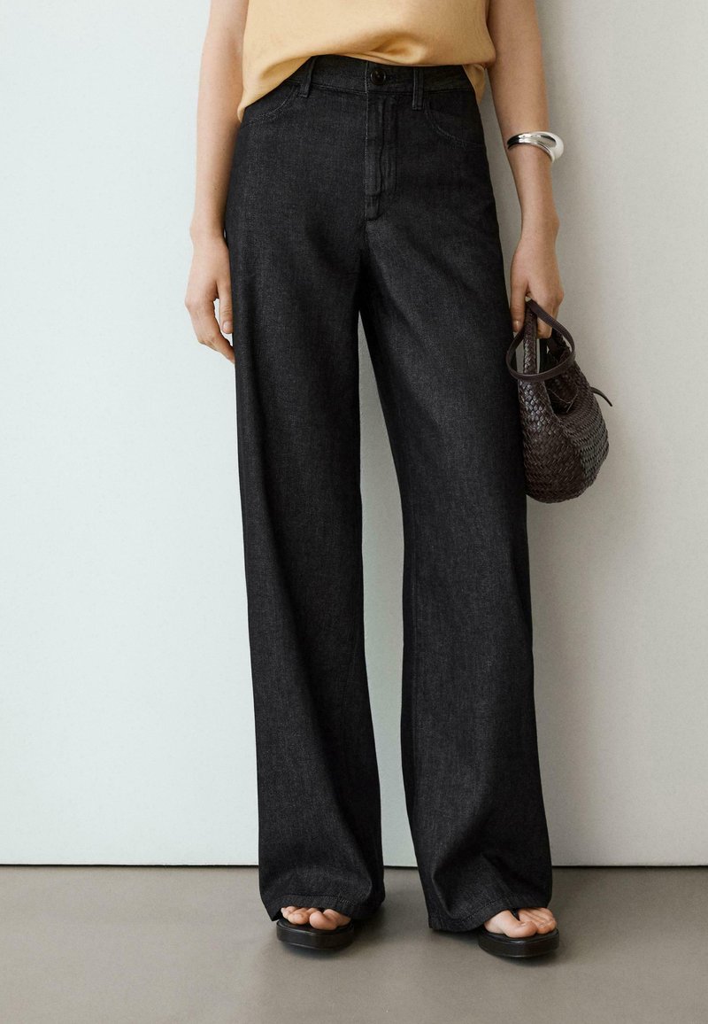 Frau trägt schwarze Jeans mit weitem Bein, beiges Oberteil, schwarze Sandalen, silbernes Armband, hält eine kleine gewebte Handtasche an ihrer Seite.