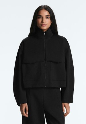 Femme aux longs cheveux foncés portant une veste noire zippée avec des poches frontales et un pantalon noir assorti, se tenant devant un fond clair.