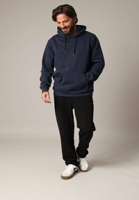 Marineblauwe hoodie met trekkoorden, voorzak en geribde onderkant; gecombineerd met zwarte broek en witte sneakers. Glad materiaal met een casual pasvorm.