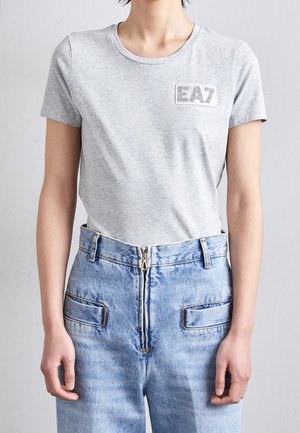 Person wearing a light gray EA7 logo t-shirt tucked into high-waisted light blue jeans with front zipper and pockets.
