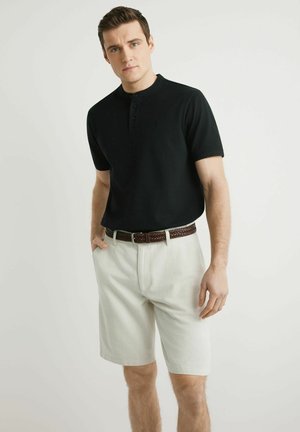 Młody mężczyzna w czarnej koszulce henley z krótkim rękawem, beżowych szortach z plecionym brązowym paskiem, stoi z rękami w kieszeniach na jednolitym tle.