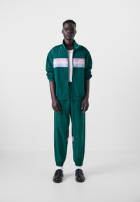 Casaco de desporto verde com riscas rosas e roxas, calças de treino a condizer, tecido texturizado e punhos elásticos. Usado com uma camisa branca e sapatos pretos.