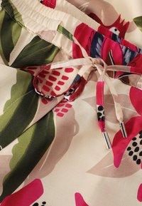 Close-up van stof met een bloemenprint in roze, groen en beige, met een gestrikte trekkoord met metalen tips op een elastische tailleband.
