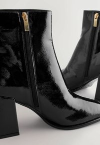 Black patent leather ankle boots with square heels, featuring gold zippers on the outer sides and a sleek, pointed toe design.