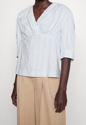 Jasnoniebieska bluzka w paski z dekoltem w serek i rękawami do połowy przedramienia, zestawiona z beżowymi, szerokimi spodniami z wysokim stanem na ciemnoskórej osobie.