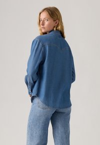 Blaue Denim-Bluse mit Rückenpasse, langen Ärmeln und Knopfmanschetten, mit einem lockeren Schnitt und leicht abgerundetem Saum.