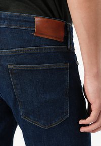Jeans en denim bleu foncé avec une texture lisse. Dotés d'une poche arrière cousue, d'un patch en cuir avec logo et de coutures jaunes contrastantes.
