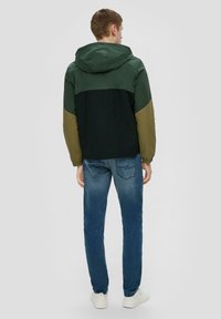 De kleurblok-jack heeft een donkergroene bovenkant, een zwarte onderkant en olijfgroene mouwen. Gemaakt van lichtgewicht materiaal met een capuchon. Gecombineerd met blauwe jeans.