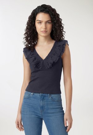 Donna con capelli ricci e scuri che indossa una canotta blu navy senza maniche con scollo a V arricciato e jeans blu, in piedi davanti a uno sfondo chiaro uniforme.