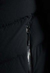 Détail d'une doudoune noire avec une fermeture éclair brillante, un tissu lisse, une texture matelassée et un design de couture en biais pour plus de style.