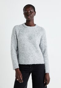 Pull en peluche gris clair avec un col rond, des manches longues et une coupe décontractée. La texture du tissu est douce et légèrement côtelée en bas.