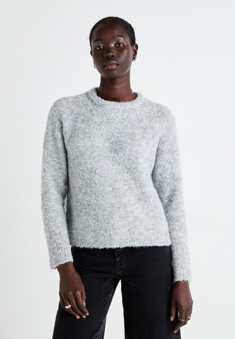 Pull en peluche gris clair avec un col rond, des manches longues et une coupe décontractée. La texture du tissu est douce et légèrement côtelée en bas.