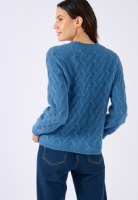 Blauwe gebreide trui met verhoogde kabeltextuur, ronde halslijn en lange mouwen, gecombineerd met donkerdenim jeans.