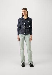 Mørk denimjakke med belte i livet, med krage, sølvknapper og to brystlommer. Parret med lyseblå jeans og svarte sko.