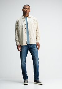 Beige, knoop-up shirt met twee voorzakken, gelaagd over een lichtblauw T-shirt en gecombineerd met donkerblauwe jeans en zwart-witte sneakers.