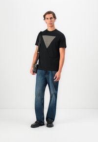 Camiseta negra con un patrón geométrico beige en el pecho, combinada con jeans de mezclilla azul y zapatos negros, sosteniendo un pequeño bolso negro.
