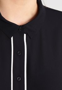Chemise noire à col avec trois boutons noirs et des rayures verticales blanches le long de la ligne des boutons sur le haut de la poitrine et le cou d'une personne.
