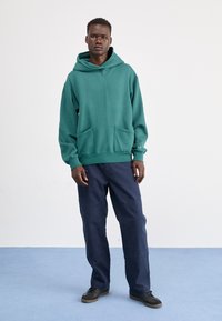 Zielona bluza z kapturem, z przednią kieszenią i sznurkiem ściągającym, w połączeniu z granatowymi spodniami. Materiał wydaje się miękki i w swobodnym stylu.