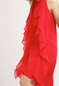Robe en chiffon rouge avec des volants en cascade sur un côté, sans manches, tissu léger et silhouette fluide.