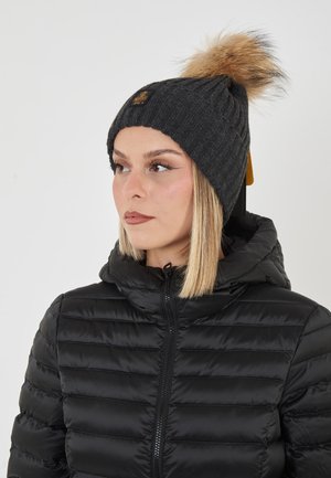 Donna che indossa un giubbotto imbottito nero e un berretto a maglia grigio scuro con un grande pom-pom di pelliccia marrone, che guarda leggermente a sinistra.