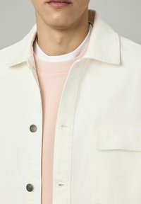 Gros plan sur un homme portant un pull en maille rose clair sous une veste en denim blanche à moitié boutonnée avec des boutons en métal visibles et une poche poitrine.