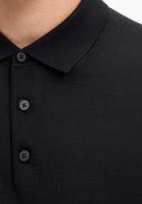 Schwarzes Poloshirt aus strukturiertem Stoff, mit einem traditionellen Kragen und einer Knopfleiste mit drei Knöpfen. Die Knöpfe sind passend und rund.