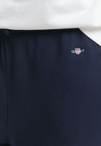 Tumesinised püksid elastse vöökohaga ja väike punase, valge ja sinise GANT logo vasakul puusal, kombineeritud valge särgiga.