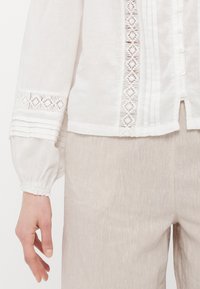 Hvit lin skjorte med broderte detaljer, teksturerte mansjetter og knappelukking. Kombinert med lysebeige, mykt teksturerte bukser.