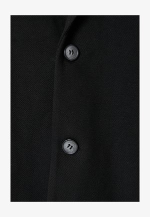 Prenda de tela negra con una textura suave, que presenta dos botones negros y un sutil patrón tejido. Diseñada con una forma clásica y ajustada.