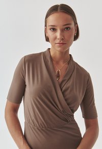 Vestido marrón de envoltura hecho de una tela suave, con mangas cortas y un detalle retorcido en la cintura. Presenta un collar de oro en capas.
