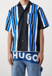 Man in een kortemouw overhemd met verticale blauwe, zwarte en witte strepen, gecombineerd met blauwe jeans waarop grote "HUGO" tekst staat.