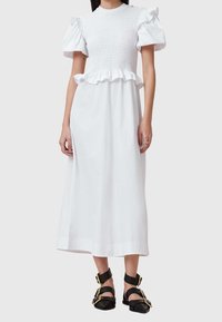 Vestido de algodón blanco con un corpiño fruncido, mangas cortas abullonadas y un dobladillo con volantes. Combinado con sandalias negras de tiras con herrajes dorados.