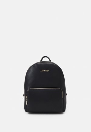 Black Calvin Klein backpack with gold logo, front zippered pocket, and top handle on a plain white background.