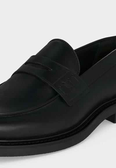 Mocassin en cuir noir avec un petit talon, comportant une bride cousue sur le devant et une finition lisse et polie.