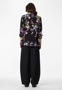 Woman standing with back to camera wearing a black floral blazer and loose black pants against a plain white background.