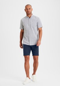 Camicia a maniche corte di colore grigio chiaro a righe, con taschino sul petto, abbinata a pantaloni corti blu scuro e scarpe da ginnastica bianche.