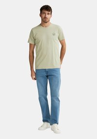 Hellgrünes Baumwoll-T-Shirt mit Rundhalsausschnitt und kleinem, bedrucktem Logo auf der Brust, kombiniert mit blauen Jeans und weißen Sneakern.