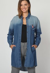 Vestido camisa de mezclilla, en degradado azul de oscuro a claro, con mangas largas, cierre de botones, bolsillo en el pecho con bordado de símbolo de paz rojo, dos bolsillos laterales.