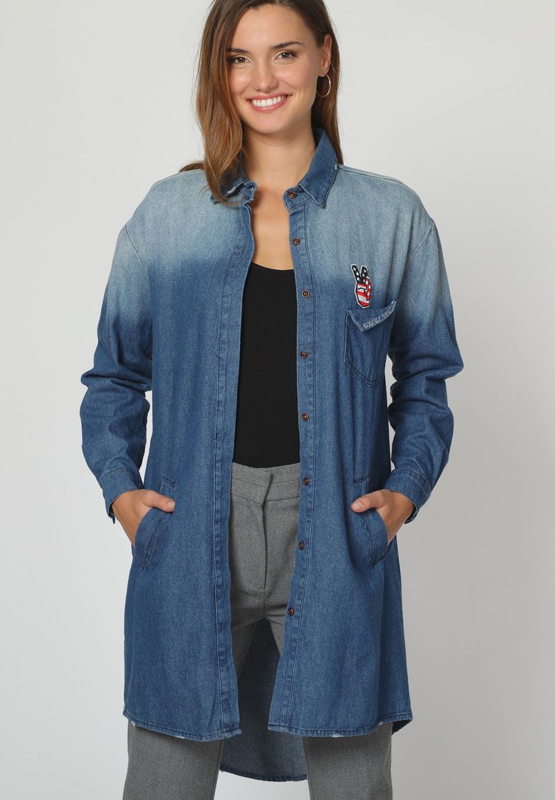 Vestido camisa de mezclilla, en degradado azul de oscuro a claro, con mangas largas, cierre de botones, bolsillo en el pecho con bordado de símbolo de paz rojo, dos bolsillos laterales.