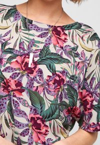 Blusa con motivo floreale su base crema chiaro, con grandi fiori rosa e viola, foglie verdi e vestibilità ampia e comoda. Collana con catenina d'argento.