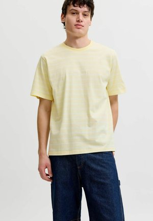 Ung mann som har på seg en lys gul stripete kortermet T-skjorte og mørkeblå jeans, står foran en ensfarget lys bakgrunn.