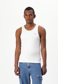 Hvit bomulls tanktop med brede stropper, rund halslinning og en liten logo på brystet, kombinert med blå denimjeans.