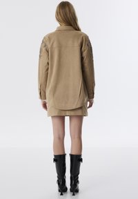 Beige suède jacket met bloemversieringen op de mouwen. Heeft een ontspannen pasvorm, afgeronde zoom en contrasterende zwarte kniehoge laarzen.