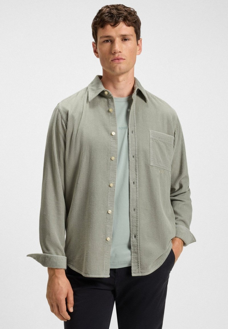 Man in een lichtgroene corduroy button-down shirt over een bijpassend t-shirt met zwarte broek, staand tegen een effen witte achtergrond.