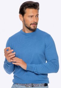 Blauer Strickpullover mit Rundhalsausschnitt, langen Ärmeln und gerippten Bündchen. Kombiniert mit einem gemusterten Gürtel und grauen Hosen.