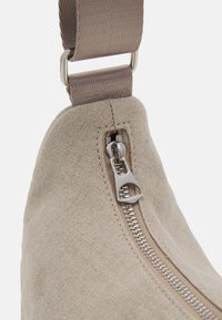 Sac en tissu beige avec une fermeture éclair argentée visible et une large sangle assortie attachée par une boucle en métal.
