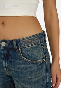 Kék farmernadrág magas derekú, látható varrással és ezüst gombbal. A derékrészen narancssárgával hímezve található a "Pegador" logó.